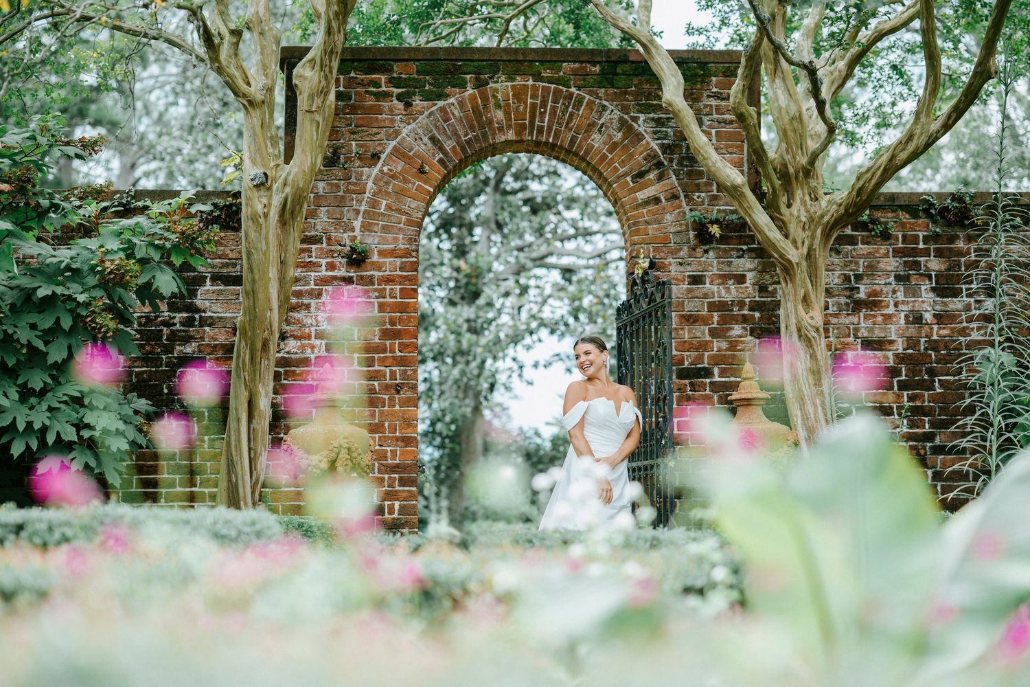 A romantic wedding couple shares a tender moment in a lush garden setting with brick archways and pink flowers.