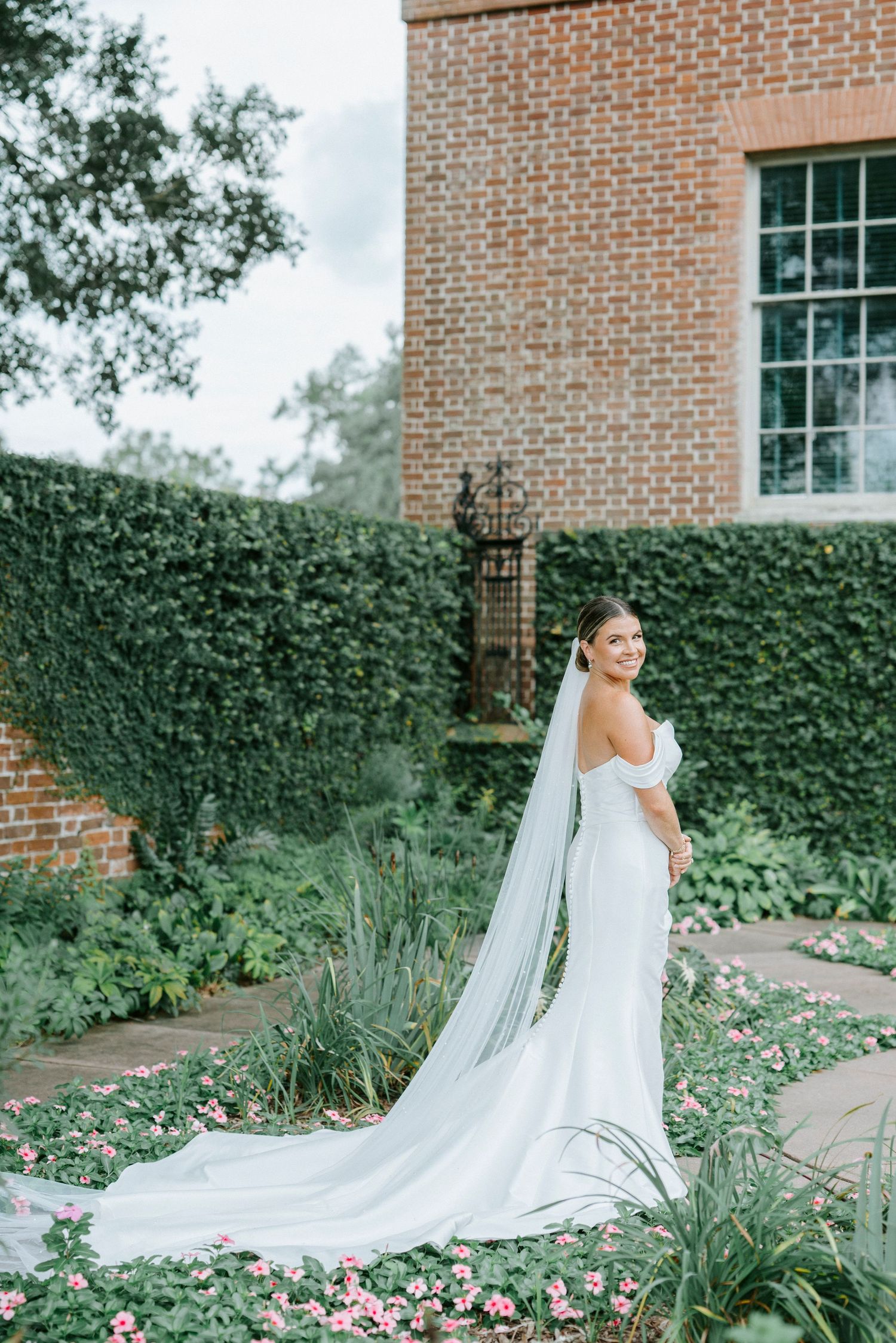 Elegant bride in white fitted gown with long flowing veil poses in garden with brick wall and ivy backdrop.