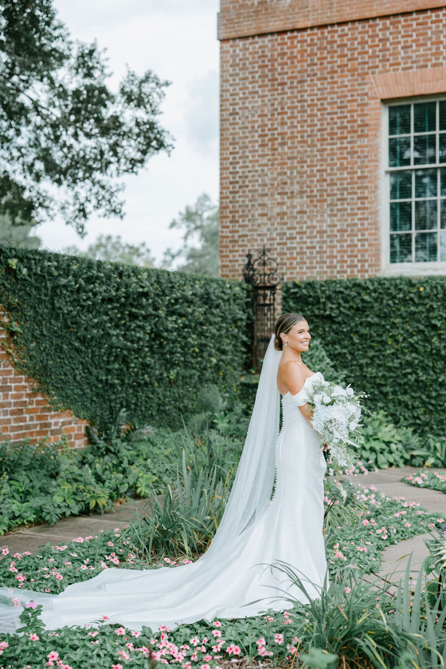 A flowing white wedding dress with a long train trails across a garden path beside a brick building and ivy wall.