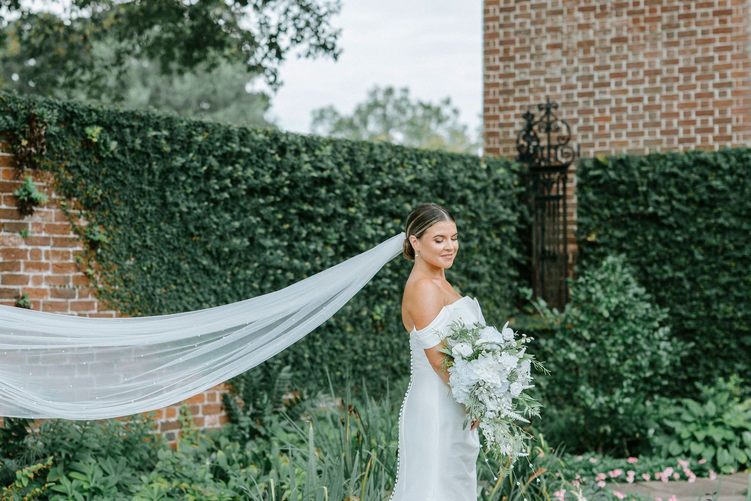 A flowing white wedding veil billows in the breeze against a backdrop of brick walls and lush green foliage.