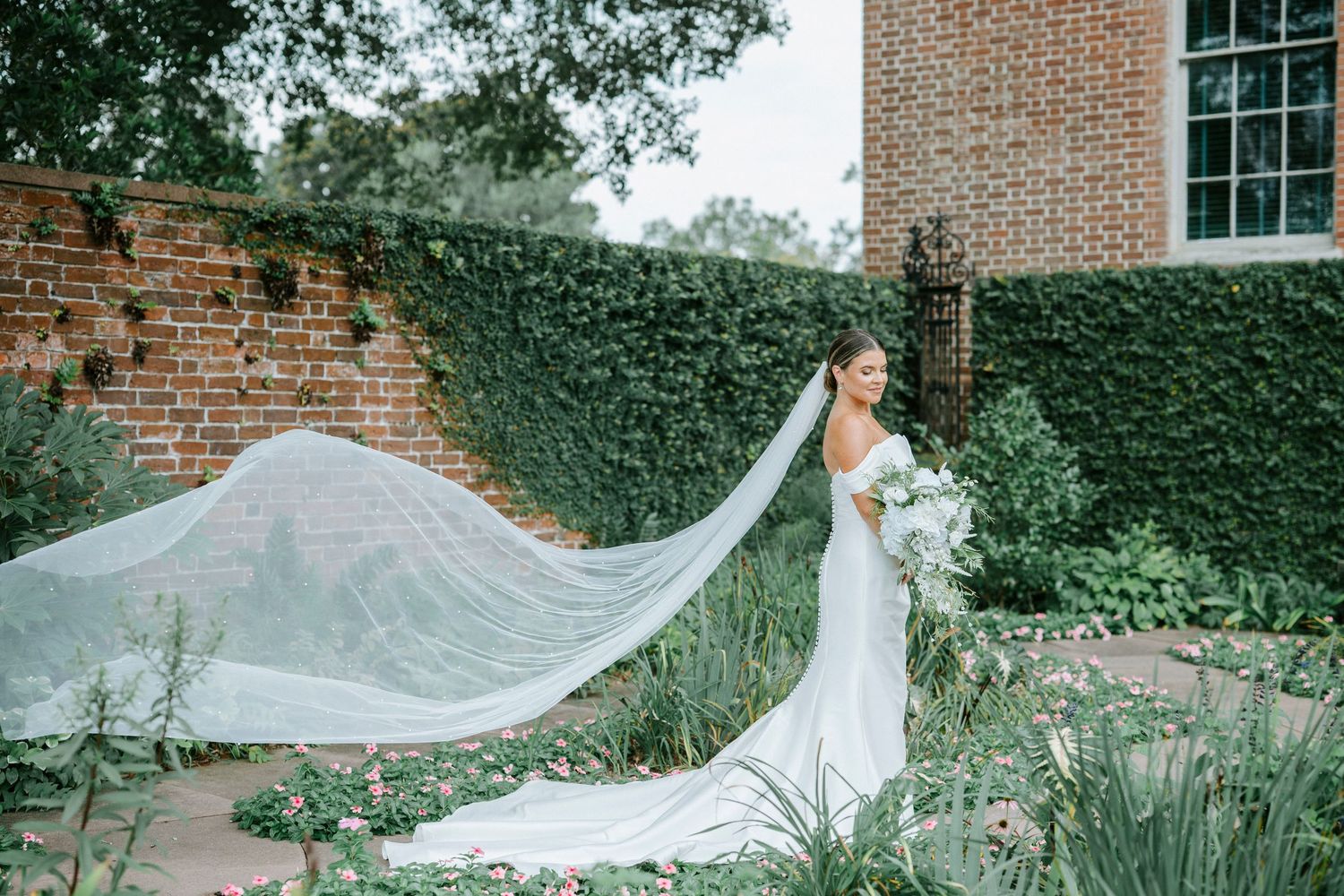 A flowing white wedding veil catches the breeze in front of an ivy-covered brick wall.