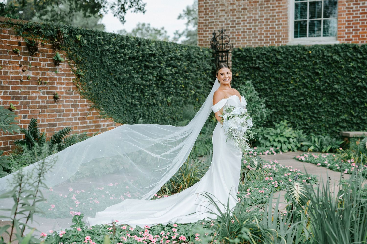 A flowing white wedding gown with long veil stands elegantly in a garden setting against an ivy-covered brick wall.