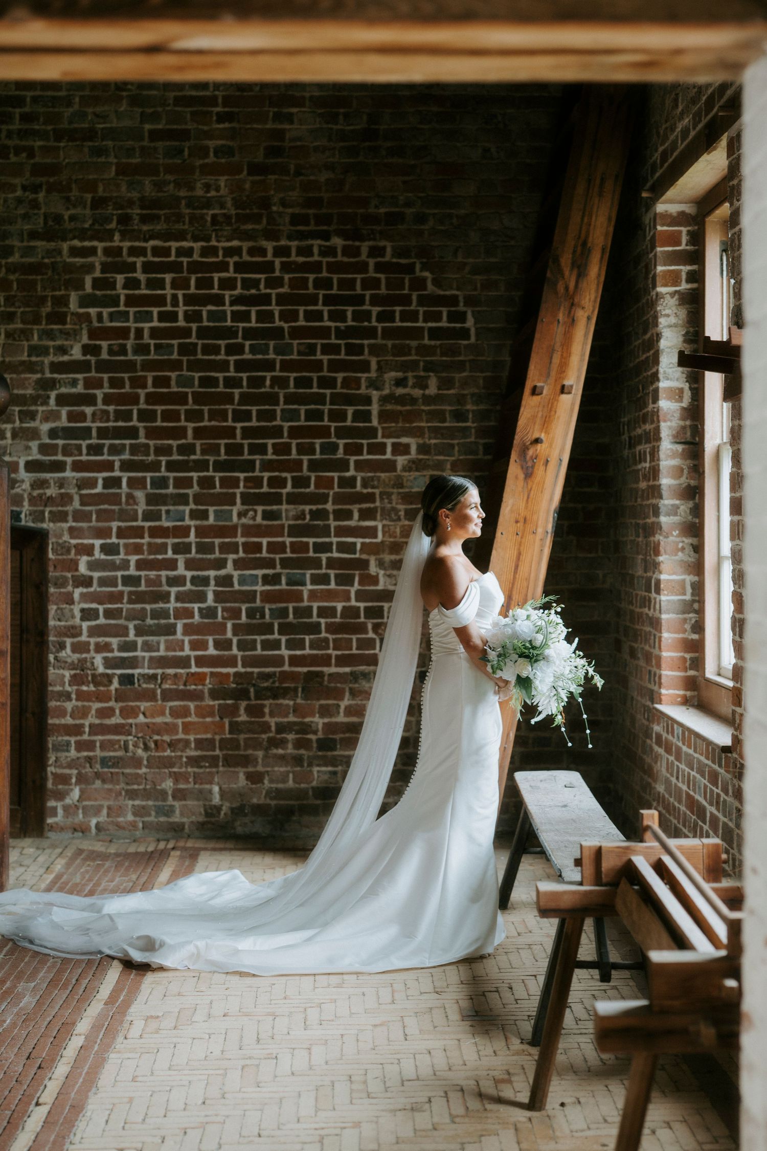 A bride in a flowing white wedding gown with a long cathedral train stands against exposed brick walls in a rustic setting.