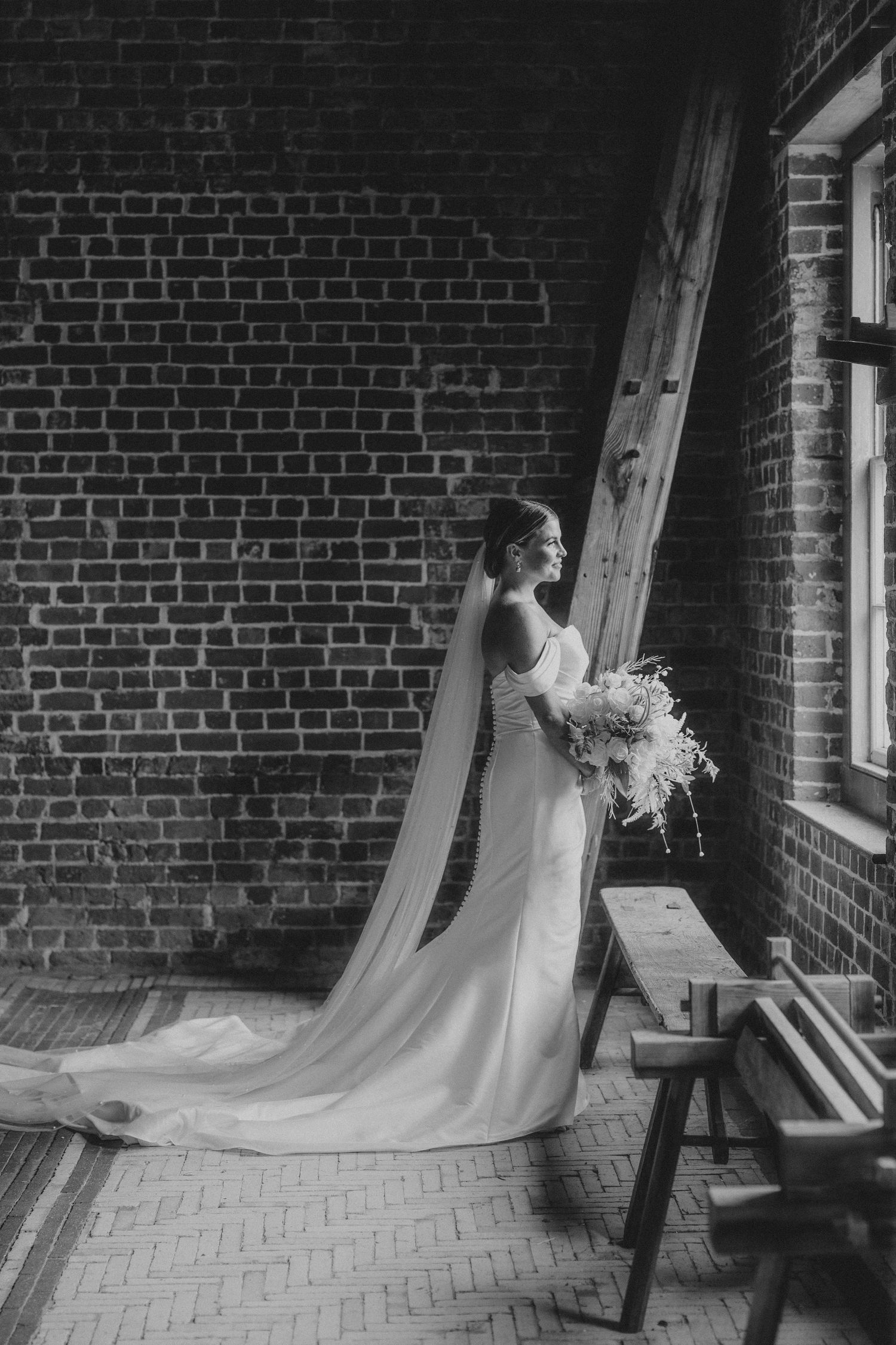 A dramatic black and white bridal portrait showcasing a long veil against exposed brick walls and wooden bench.