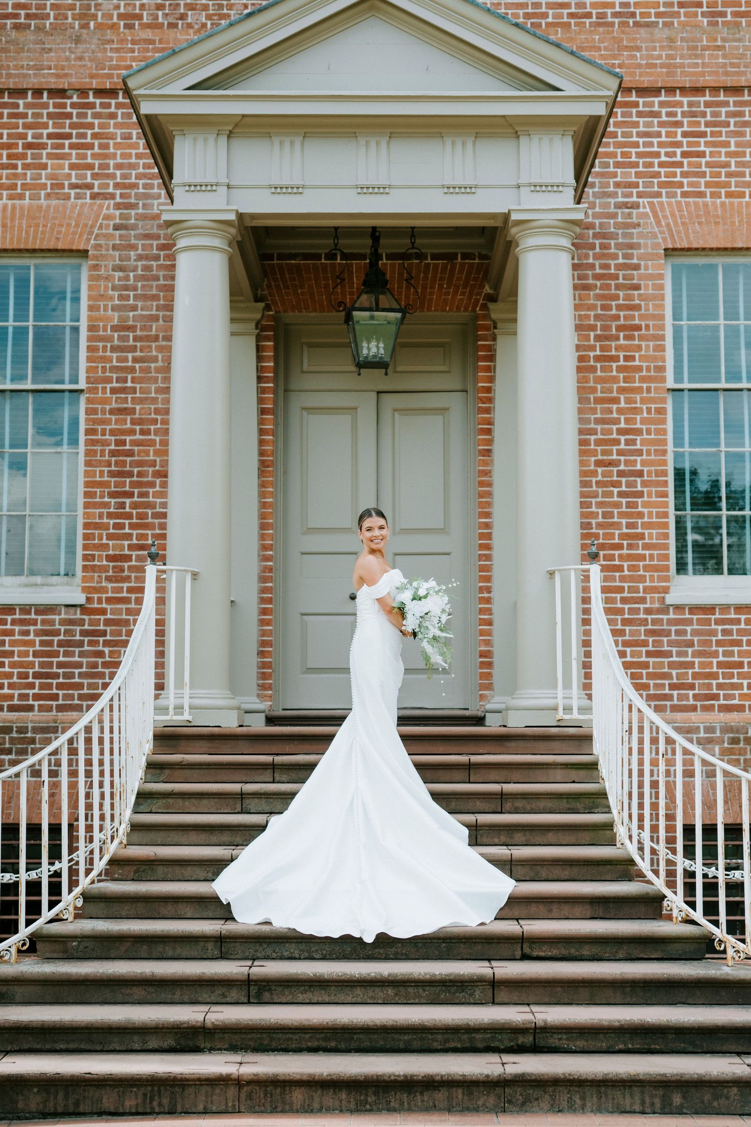 A bride in a white wedding gown poses on the brick steps of a colonial-style building with white columns and doors.