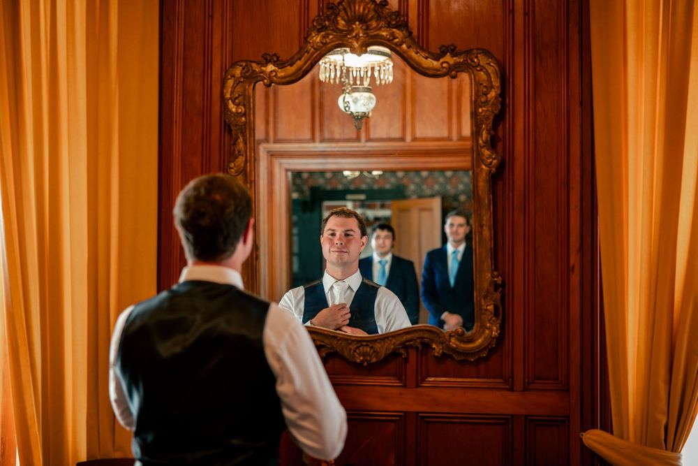 Ornate gold-framed mirror reflects a person in formal attire with orange curtains in an elegant room.
