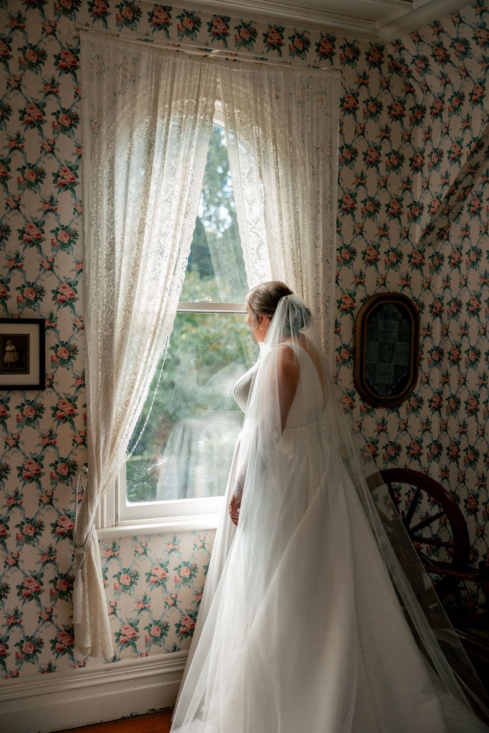 A bride in a flowing white wedding gown and veil stands by a window with floral wallpaper in a vintage-style room.