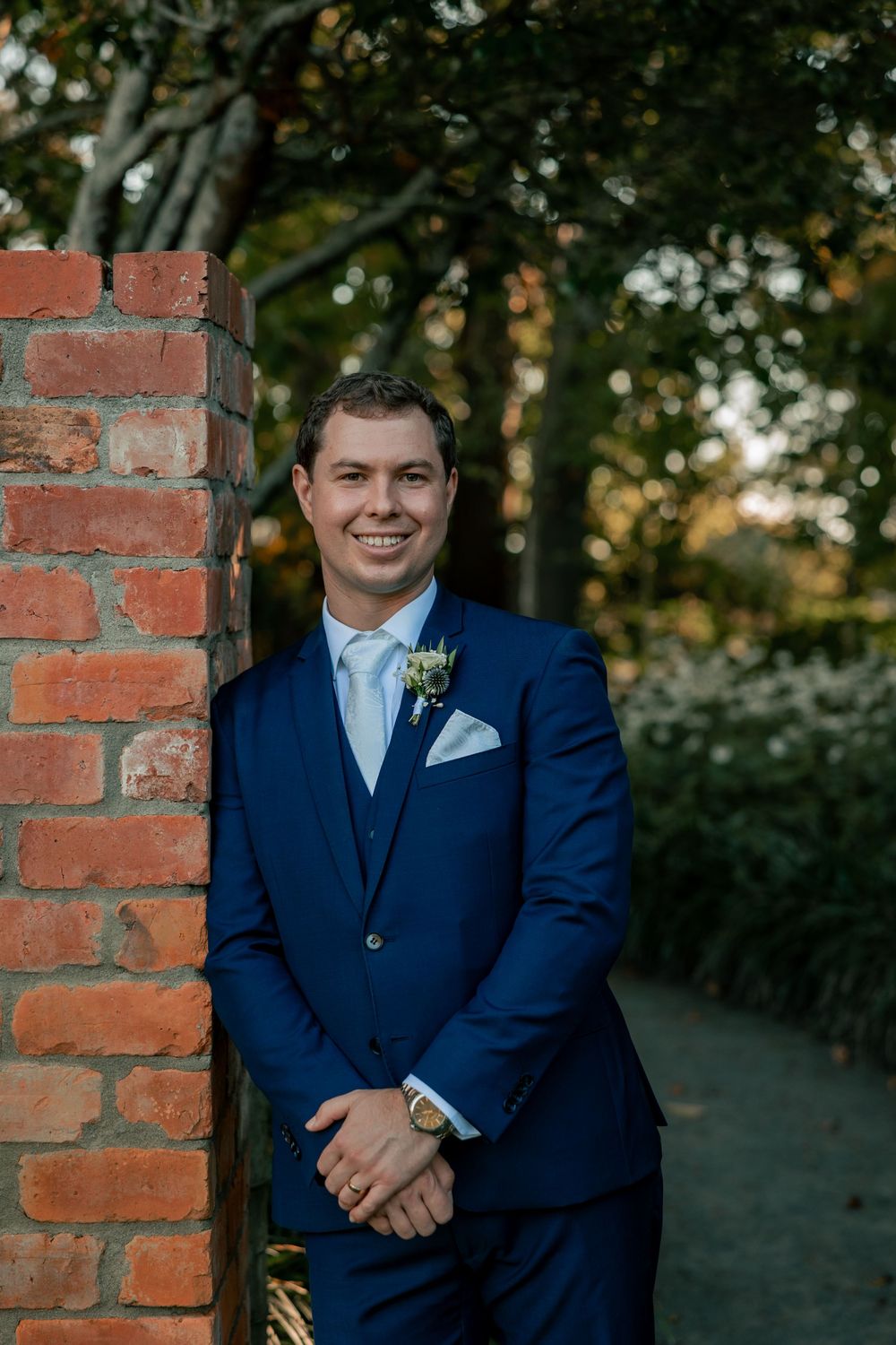 A person in a navy blue suit and white shirt stands between brick pillars in an outdoor garden setting.