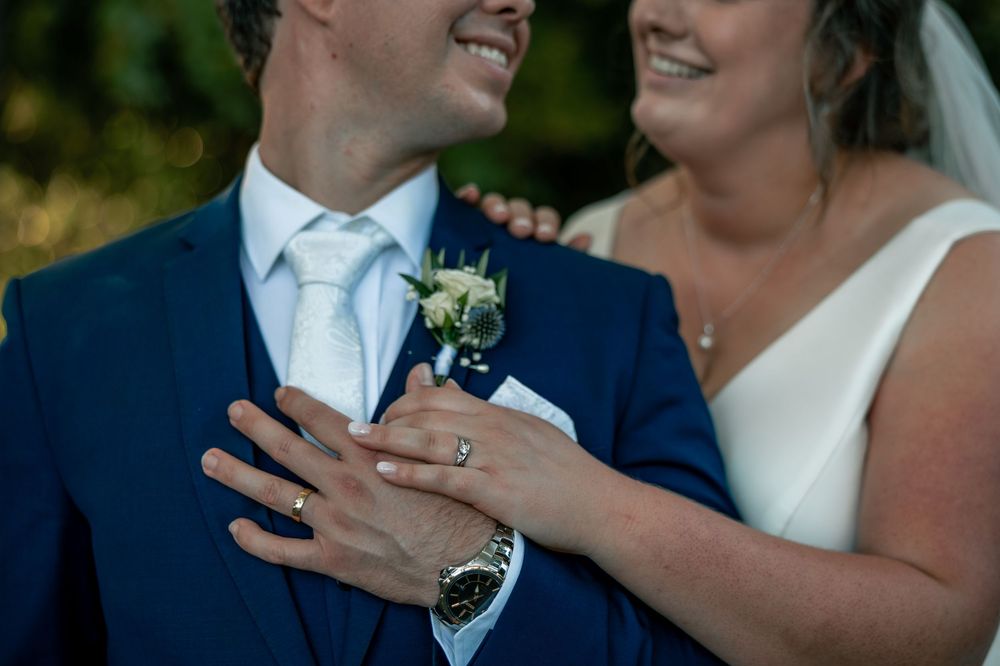 A close-up photo showing hands with wedding rings and a boutonniere against a navy blue suit and white dress.
