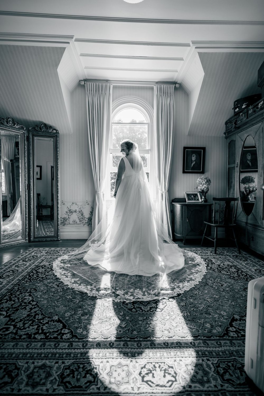 A flowing white wedding dress is illuminated by natural light streaming through windows in an elegant room with patterned carpet.