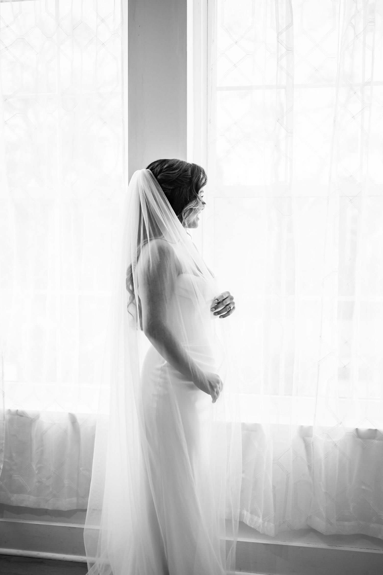 A series of ethereal black and white silhouettes show a pregnant woman in a flowing dress by a bright window.