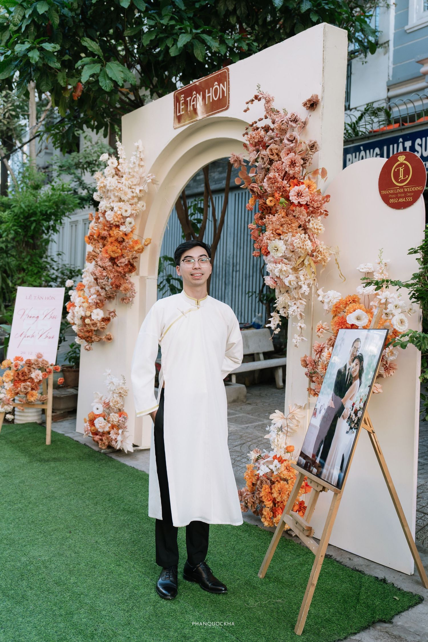 Vietnamese groom in white ao dai standing before floral arch wedding entrance with Le Tan Hon sign.