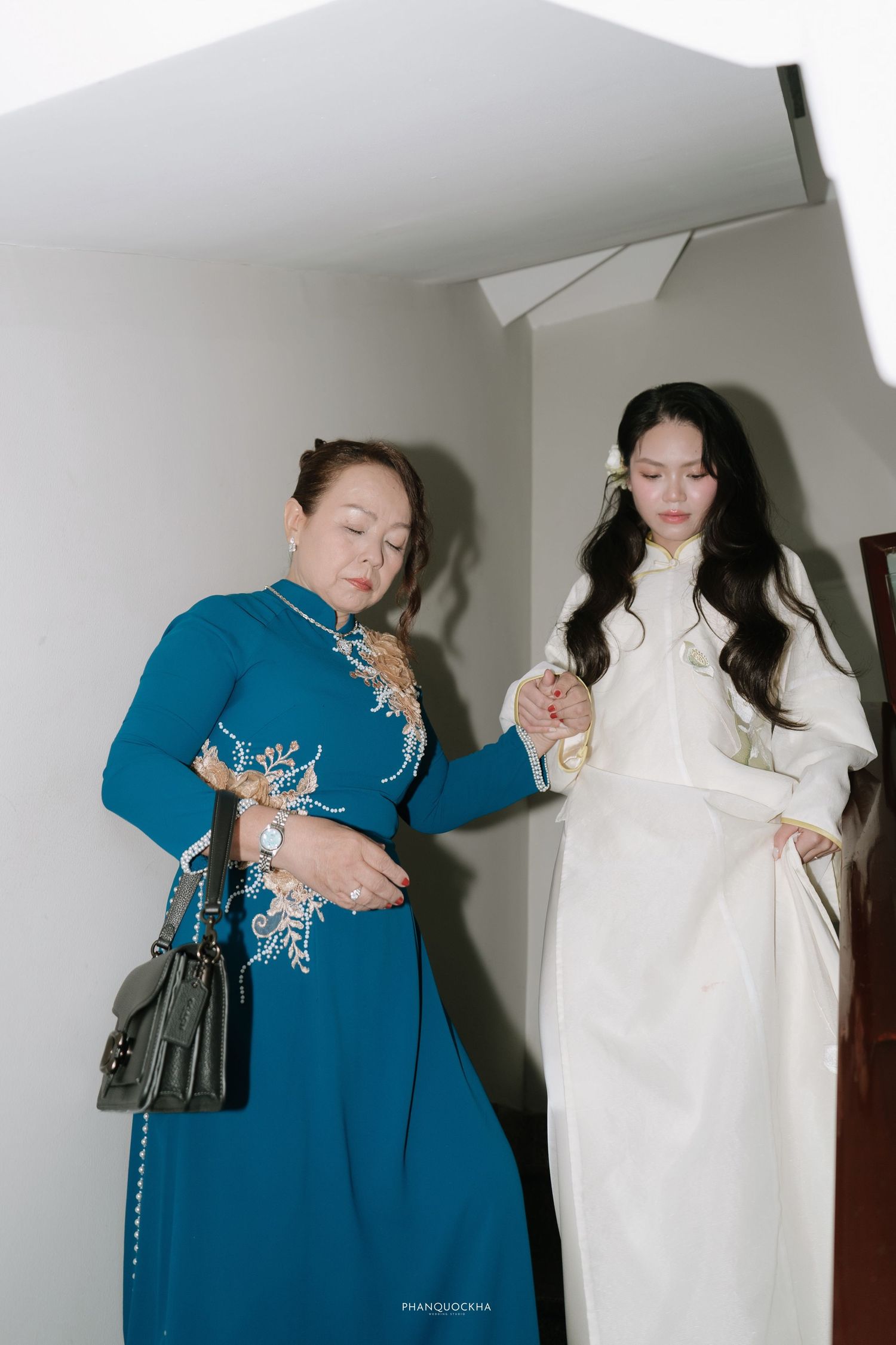 Mother in blue ao dai helping bride in white ao dai prepare before traditional Vietnamese wedding ceremony.
