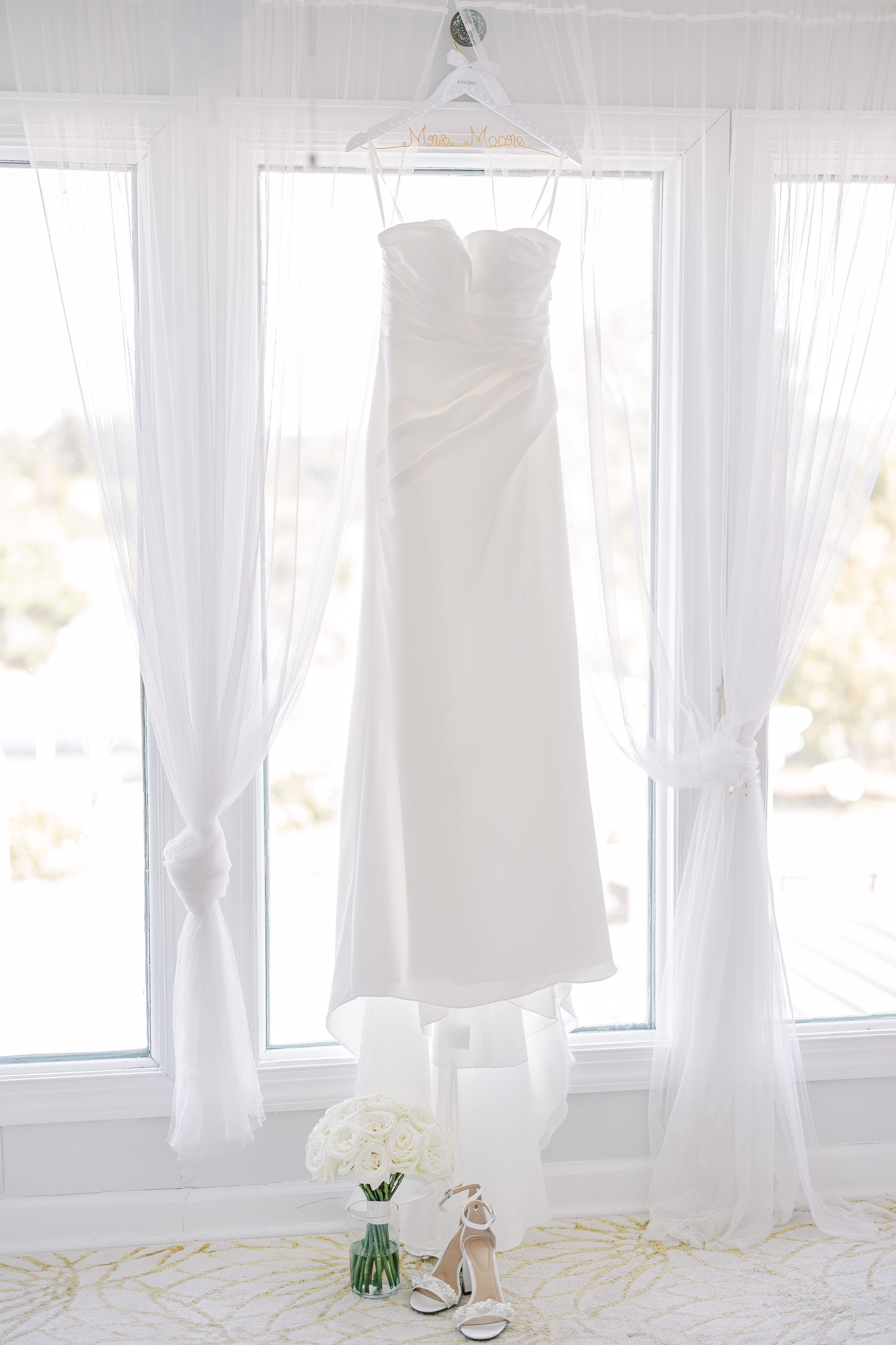White wedding dress on a hanger illuminated by natural light streaming through sheer white curtains in bright window.