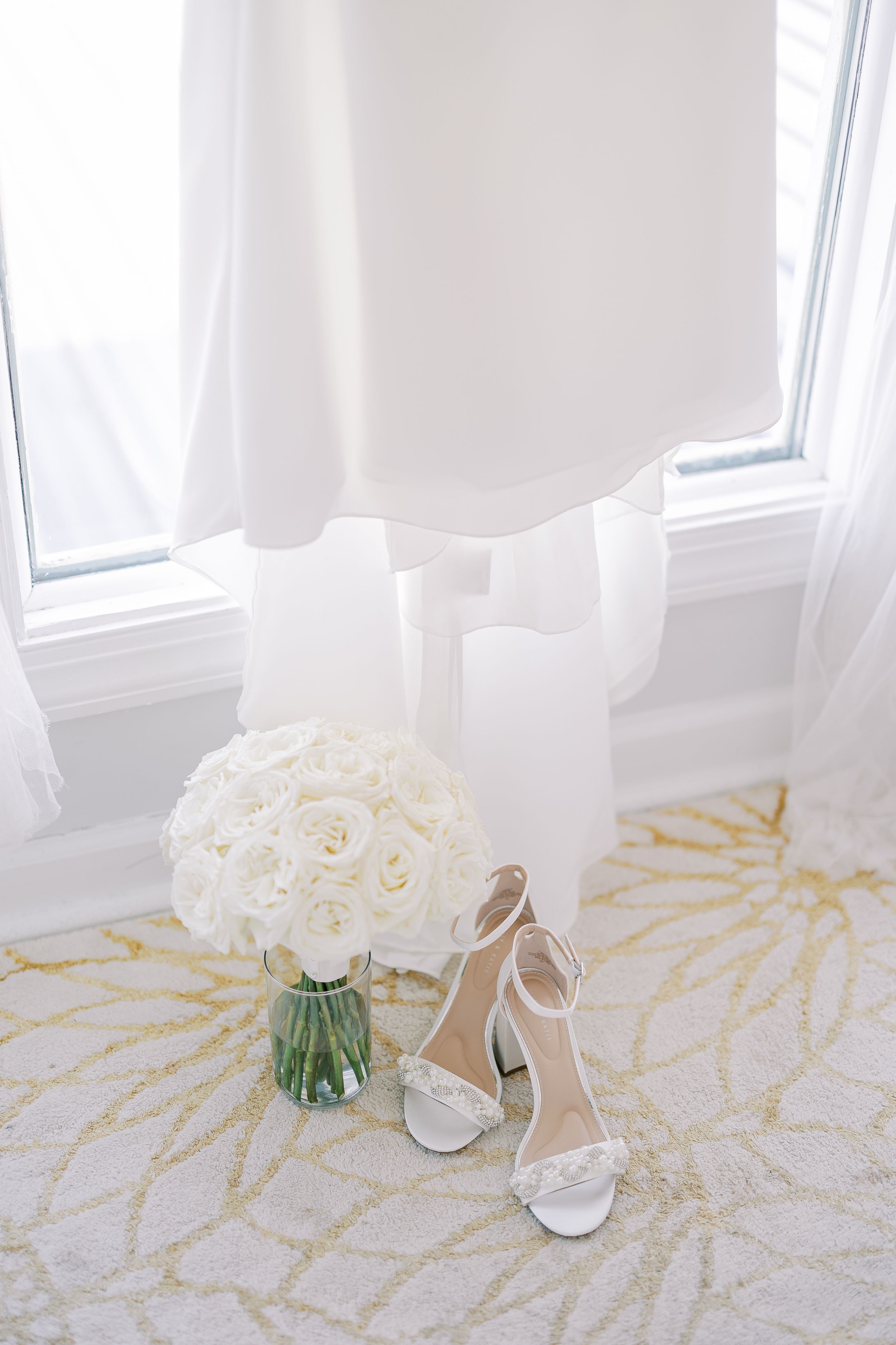 White roses in a vase sit beside elegant bridal shoes on a floor with gold geometric pattern.
