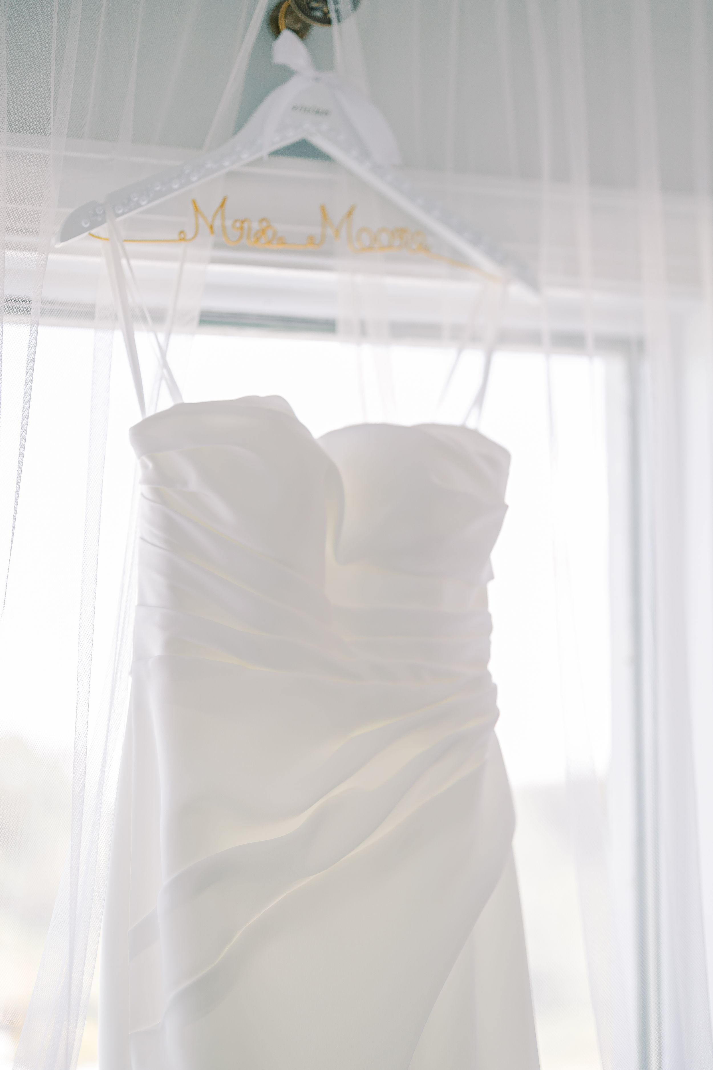 A white wedding dress hangs elegantly near bright windows in a bridal suite.