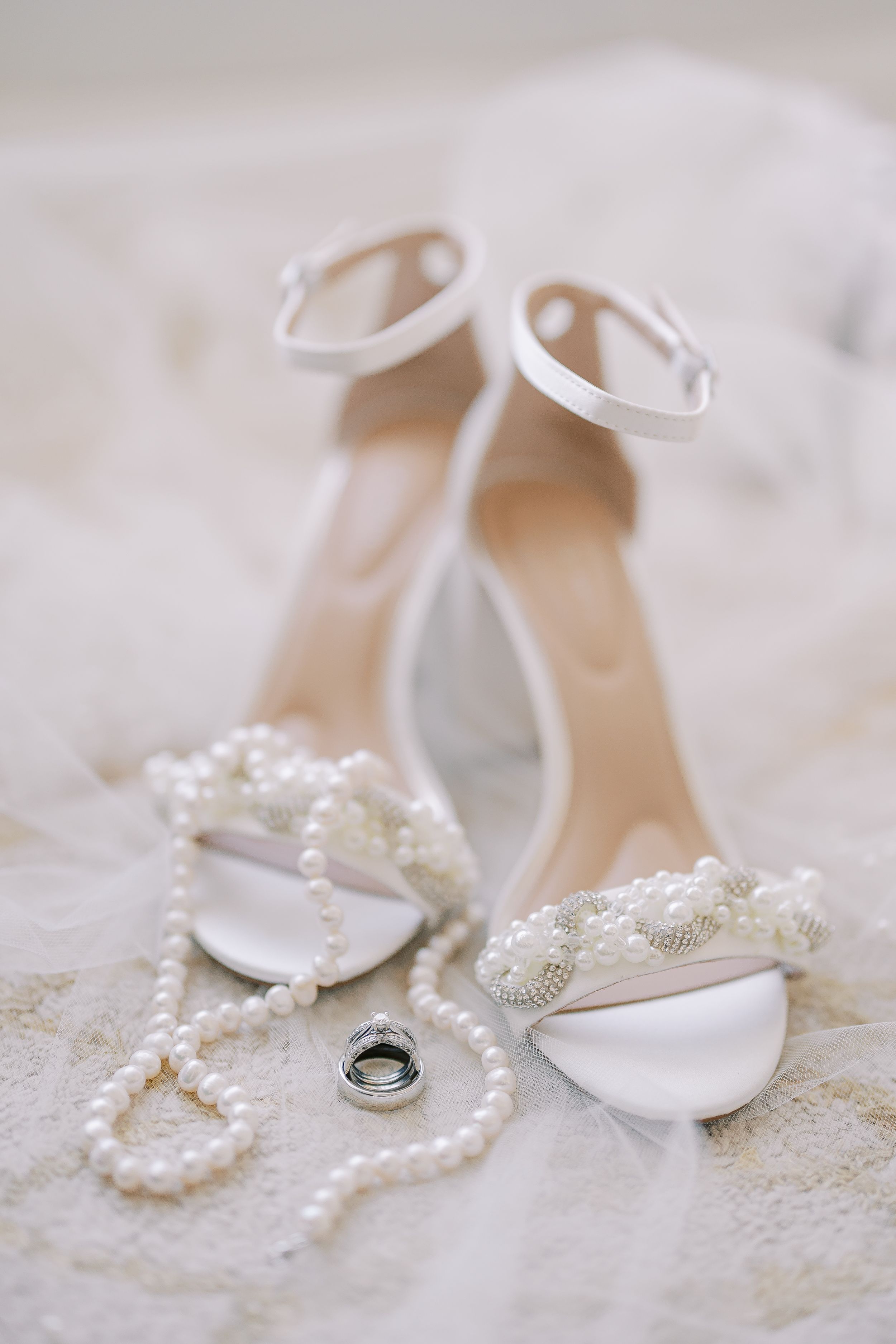 Elegant white bridal shoes with pearl details and ankle straps alongside wedding rings and a pearl necklace.