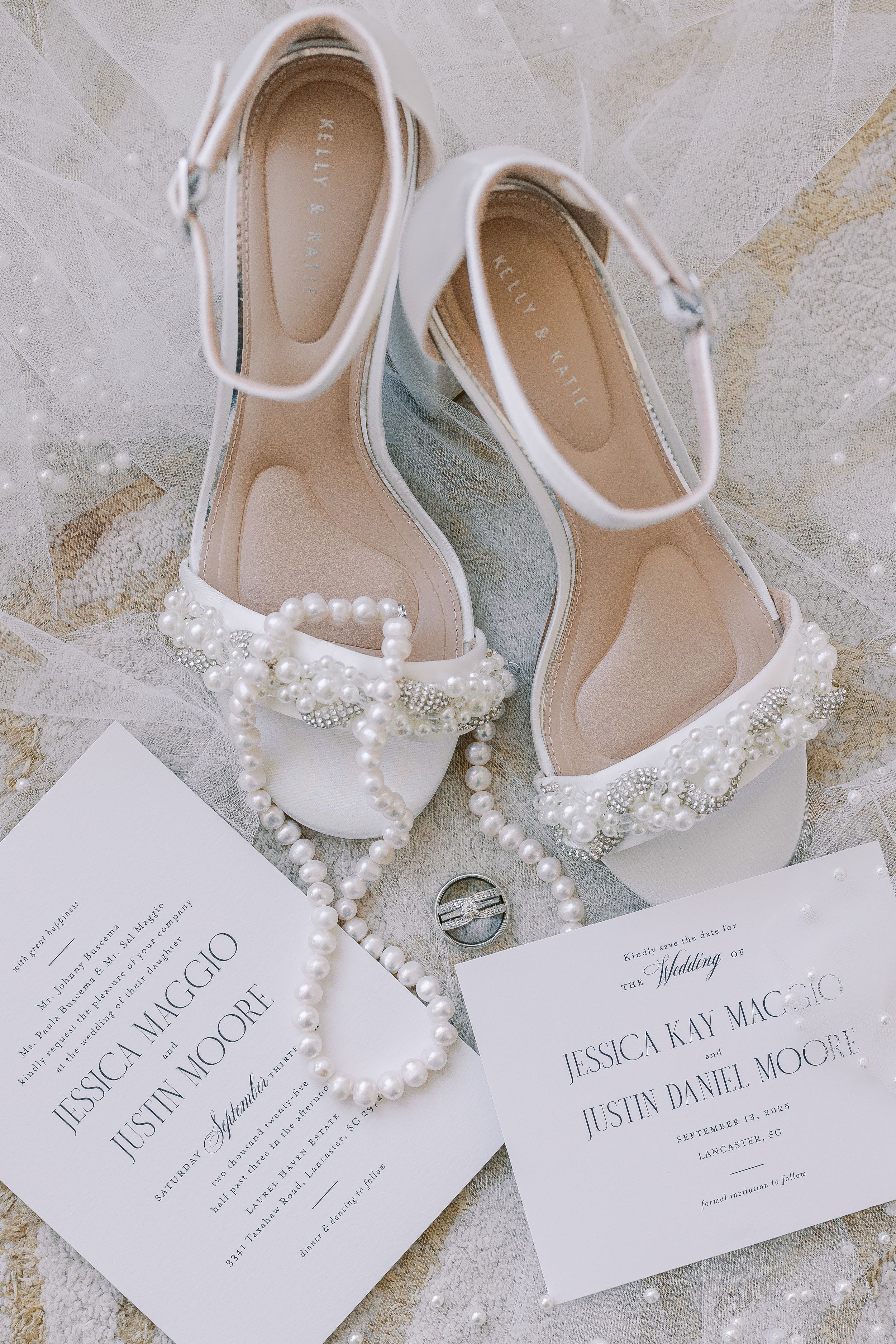 White wedding shoes with pearl embellishments displayed next to elegant invitation cards and pearl necklace on lace backdrop.