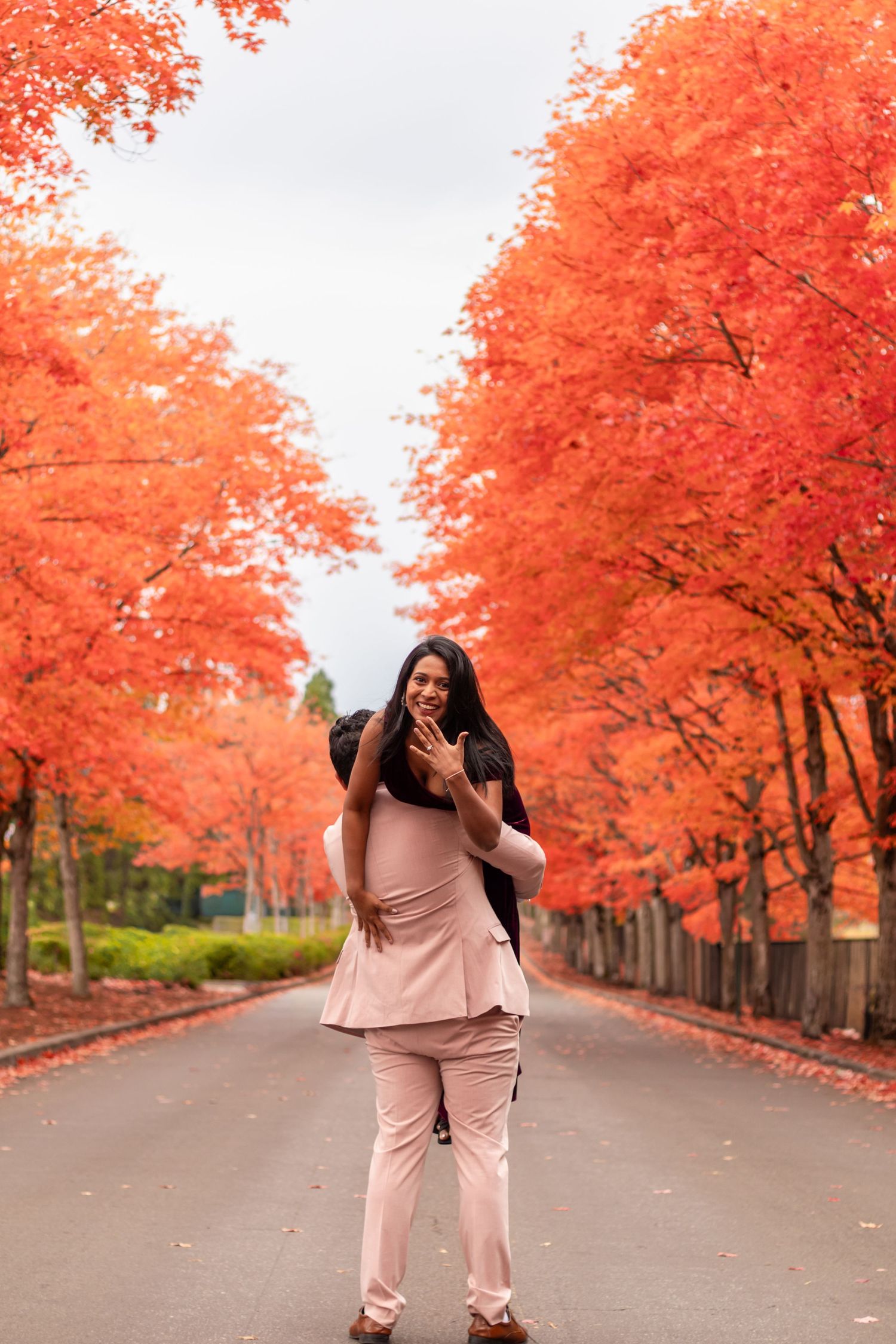 Pregnant woman in pink outfit standing on tree-lined road with vibrant red autumn foliage.