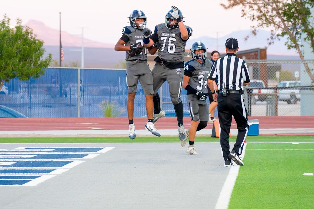 Two football players in gray uniforms celebrate a touchdown near the end zone with a referee nearby.