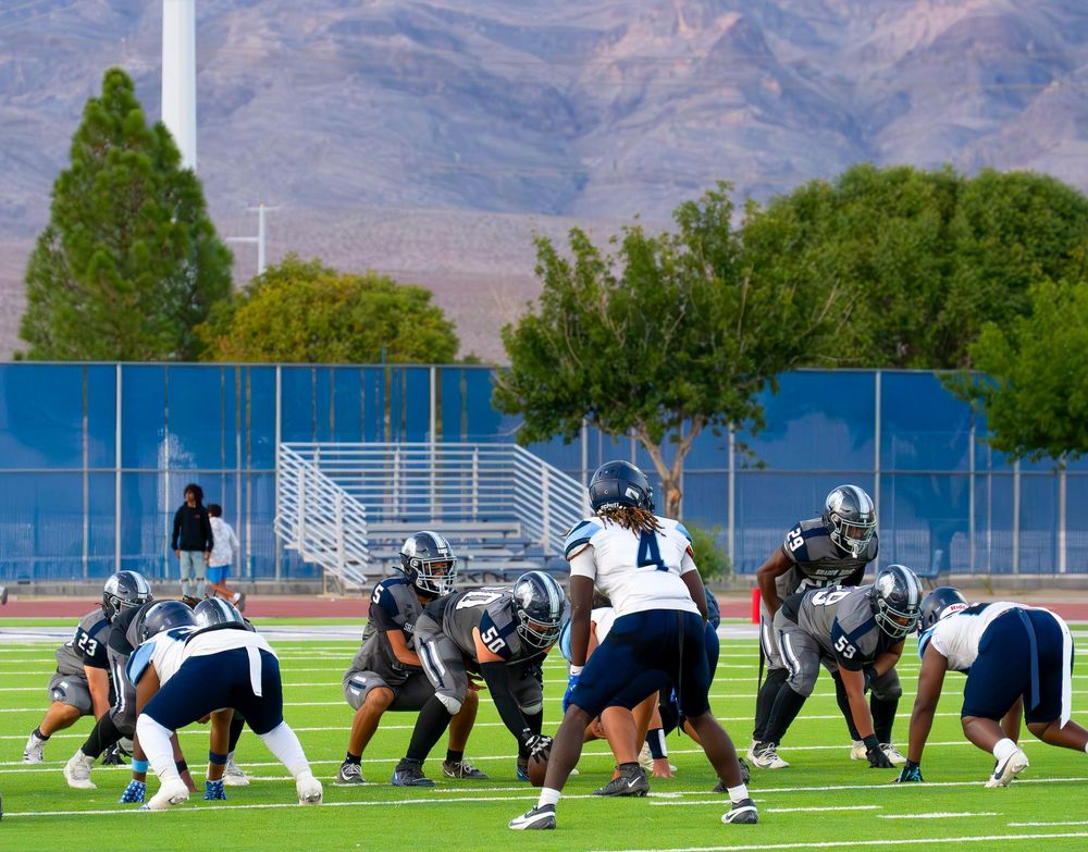 Two football teams line up at scrimmage on a blue turf field with mountains visible in the background.