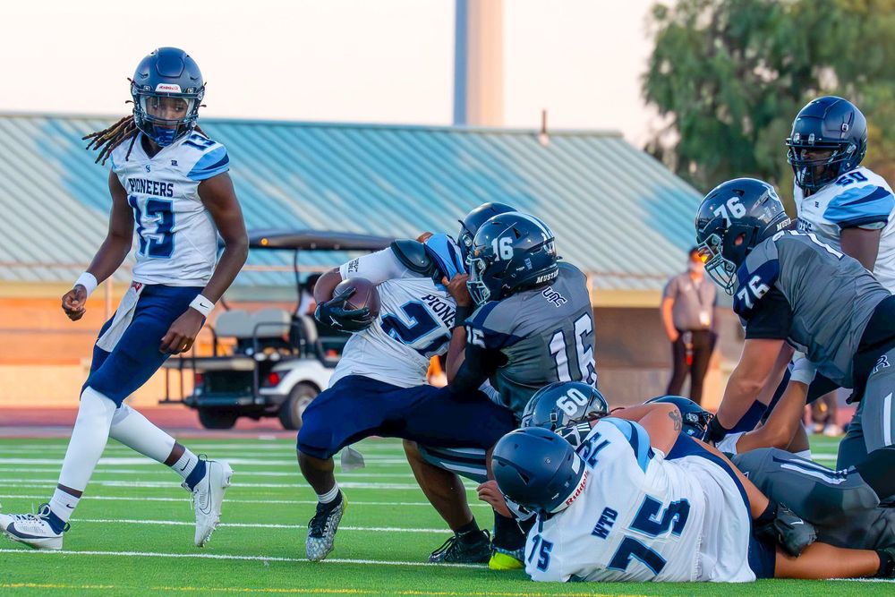 Football players in navy uniforms execute a running play while a teammate watches from the backfield.