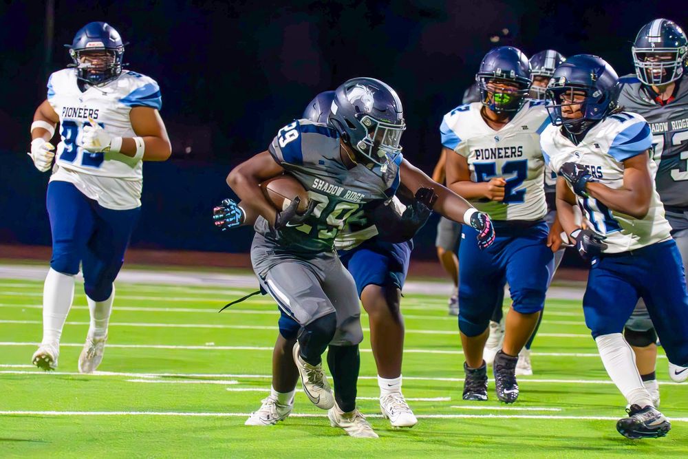 Football player in gray uniform breaks through defenders in blue Pioneers jerseys during a game.