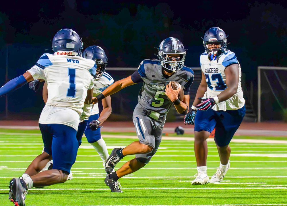 Football player number 5 in gray uniform runs with the ball past defenders in blue jerseys.