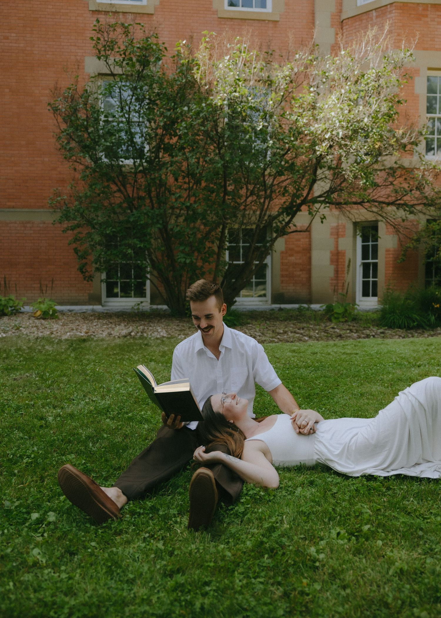 A person in a light colored shirt relaxes on a grassy lawn near a red brick building.