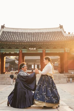 Two people wearing traditional Korean hanbok sit together in front of historic temple buildings at sunset.