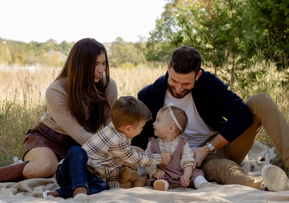 A family sits together on a blanket in a sunny field for an outdoor portrait.