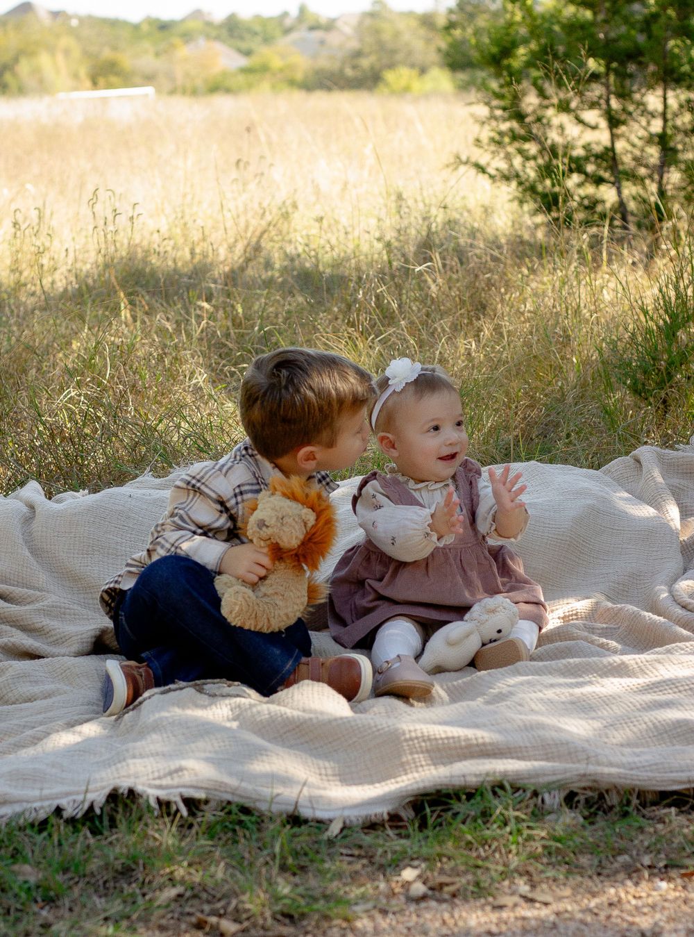Two young children smile while sitting together on a blanket in a golden field at sunset.