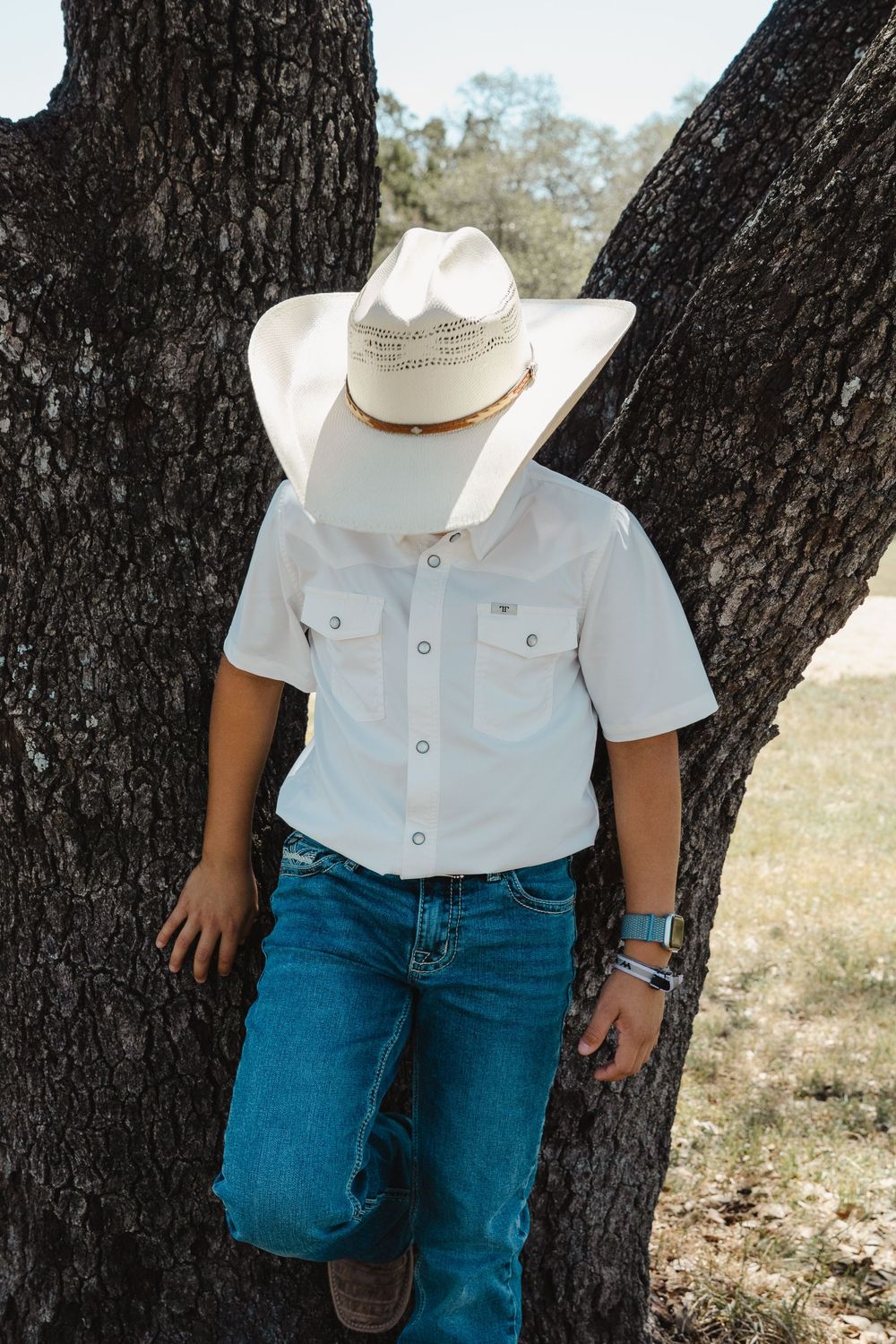 Country western style outfit with white pearl snap shirt, blue jeans and white cowboy hat leaning against a tree trunk.