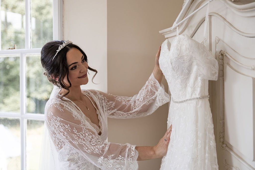 Bride in white lace wedding dress selects gown from garment rack during wedding preparations.