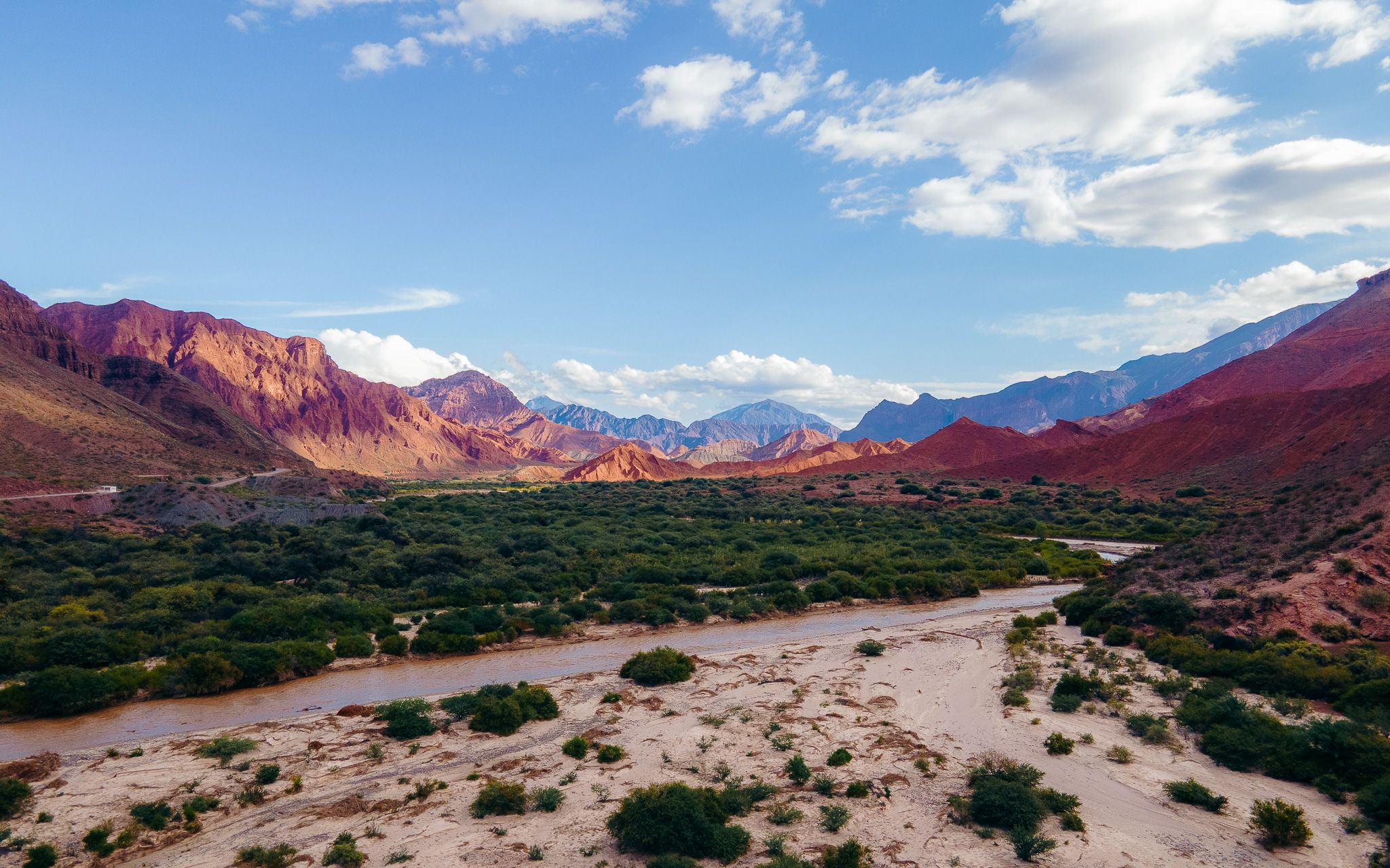 Scenic panoramic landscape of red desert rock formations and mountains under a bright blue sky with white clouds.