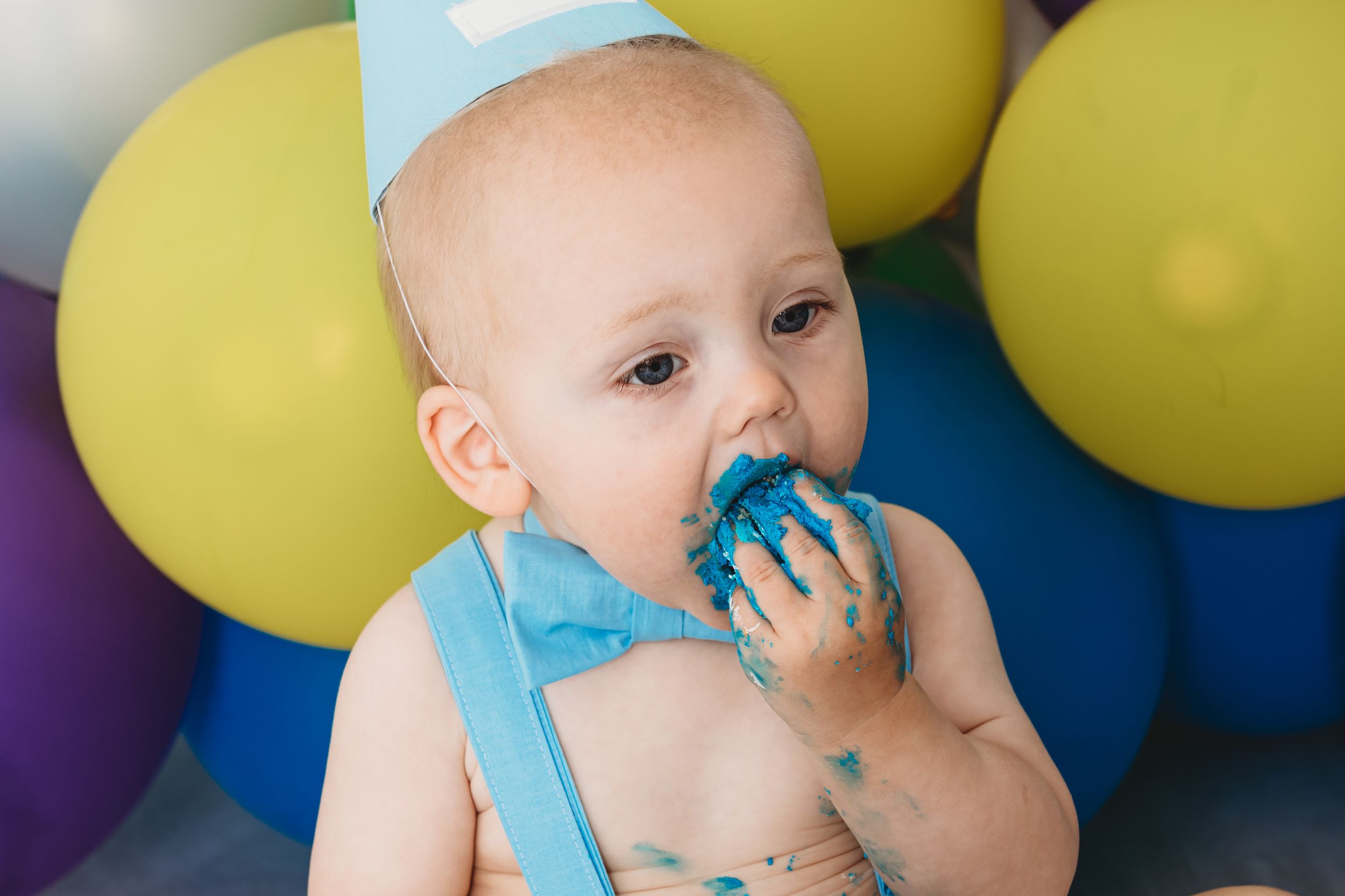 Baby in blue crown eating cake with colorful balloons in background.