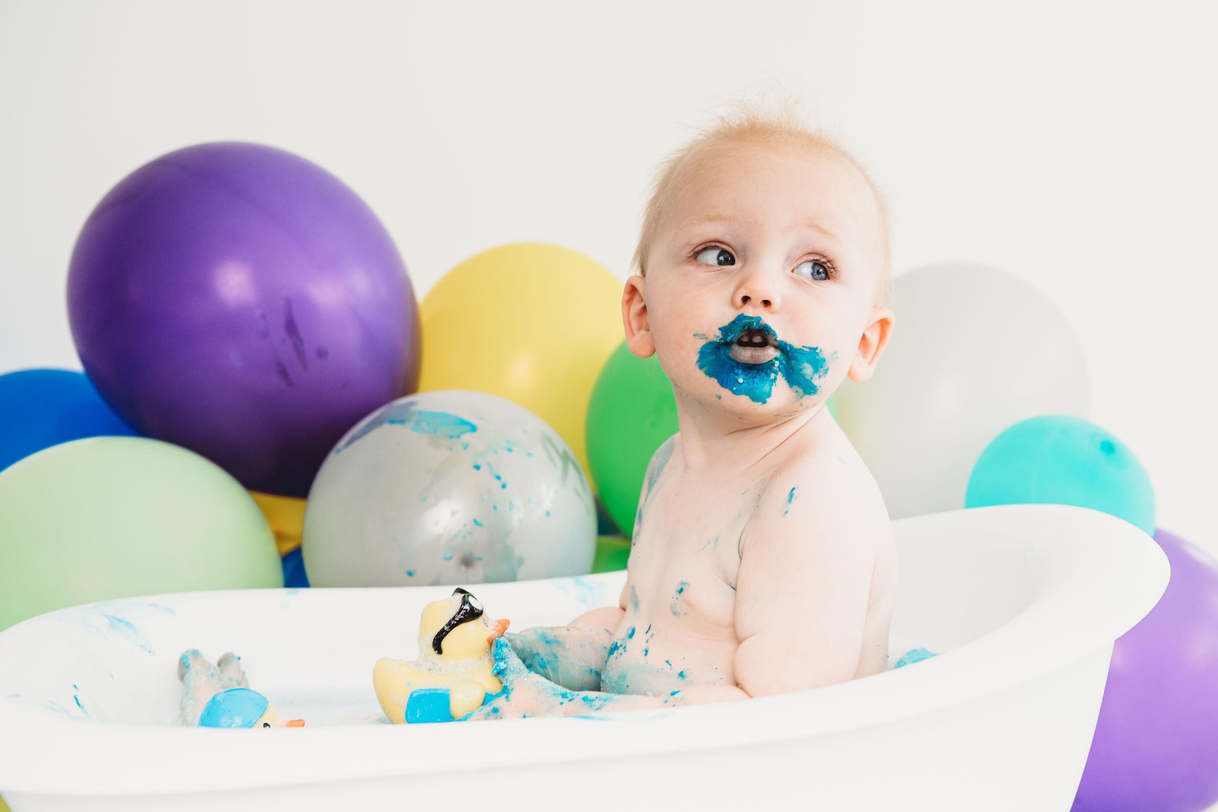 Baby in white bathtub surrounded by colorful balloons during cake smash session.
