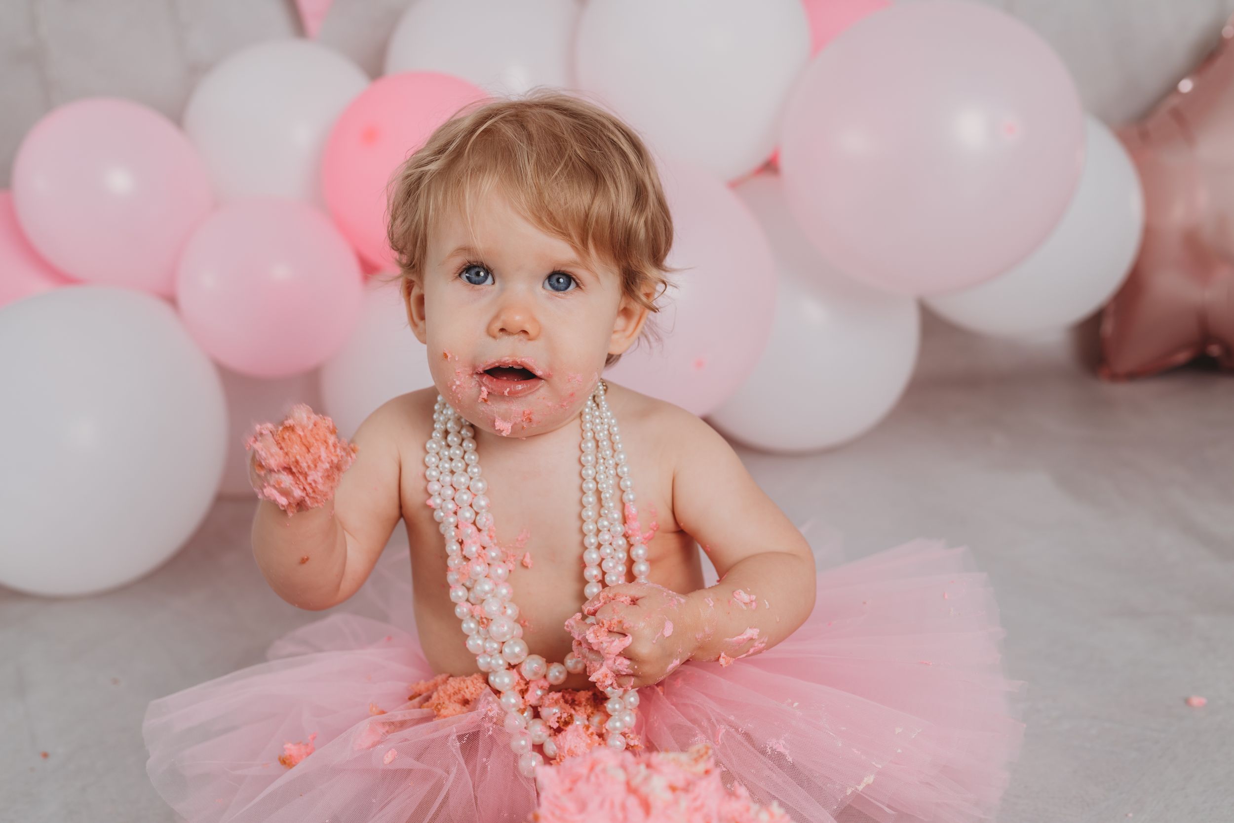 Small child in pink tutu and pearls poses with pink and white balloon backdrop.