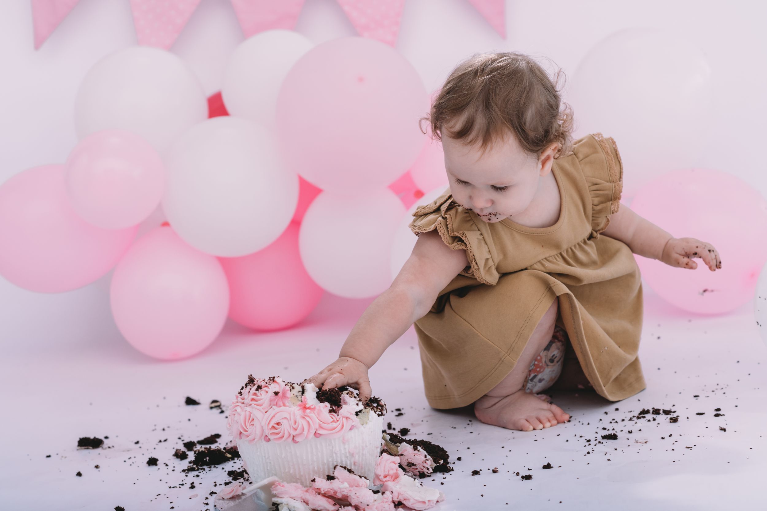 Small child in brown dress playing with cake and pink balloons during a cake smash photography session.