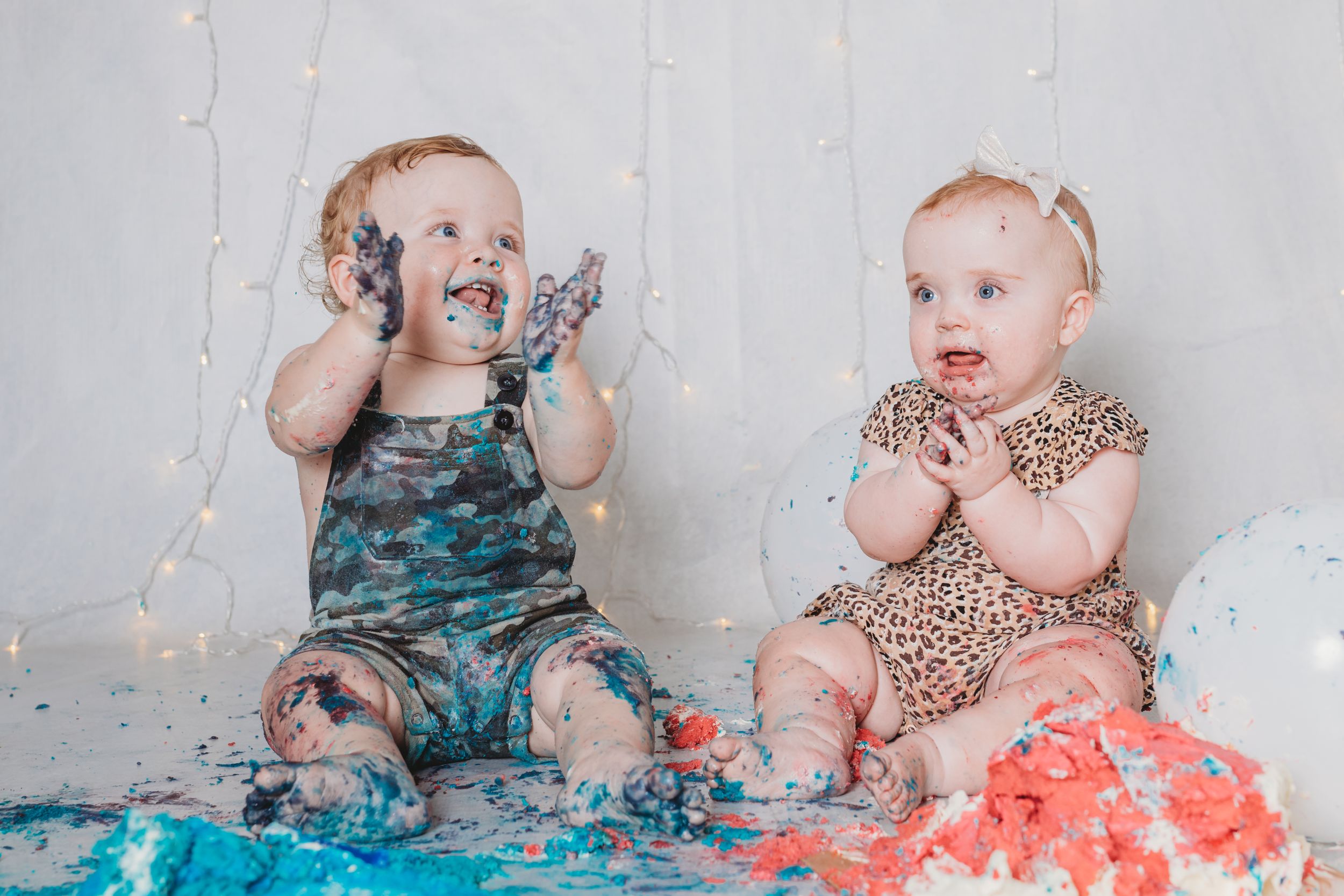 Two babies sitting together covered in blue and pink cake during cake smash photo session.