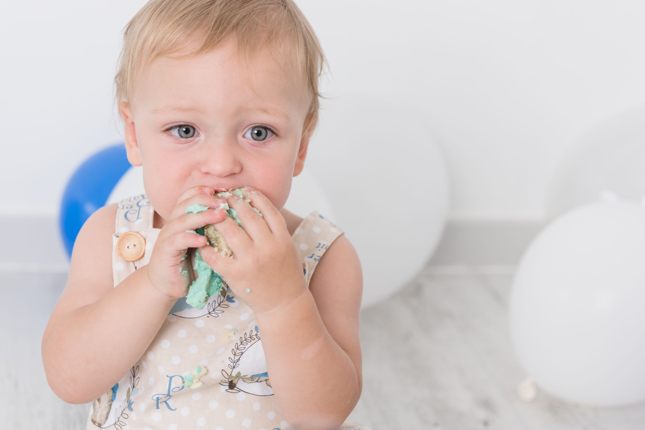 Baby in floral outfit with white balloons chewing on toy.