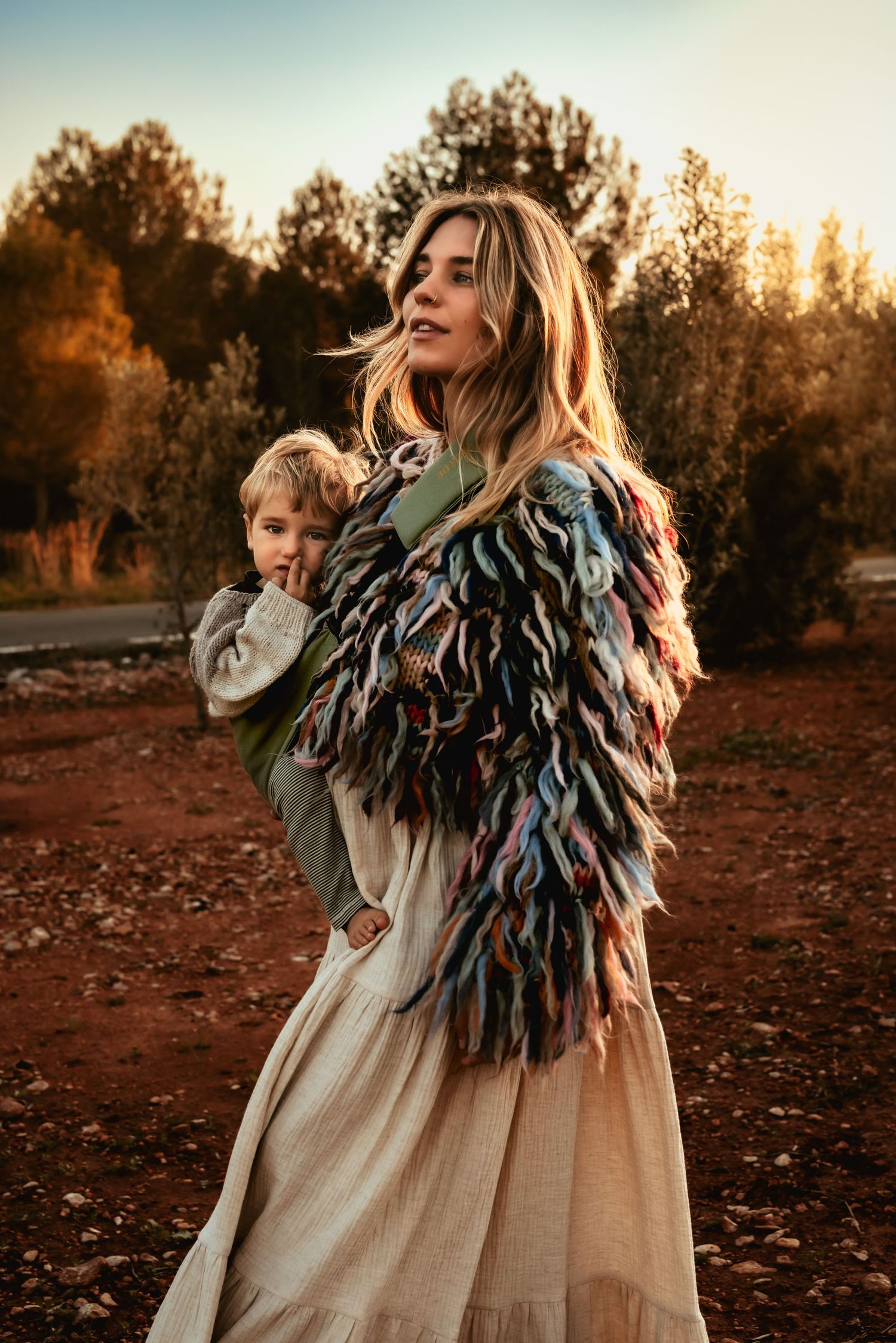 A mom in Javea  wearing a flowing dress and textured feather jacket stands in a golden autumn field at sunset. Javea maternity photographer