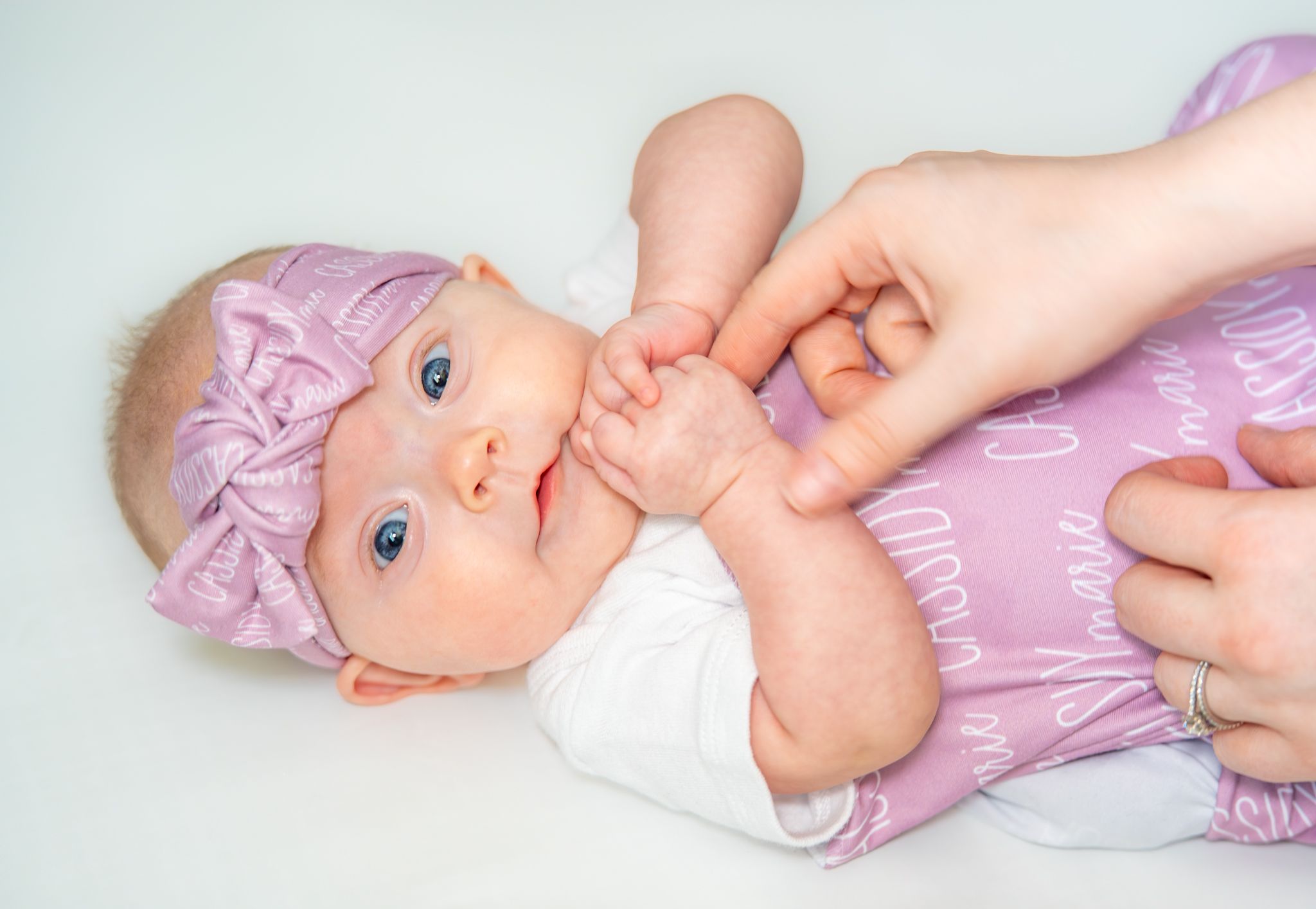 Baby in a lavender outfit and headband lying on back while someone holds their hands. Quad Cities Newborn Photography