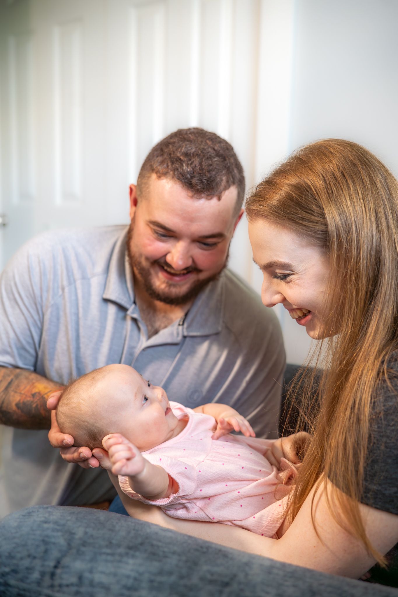 A happy couple smiles lovingly while holding and interacting with their newborn baby wearing pink. Quad Cities Newborn Photography