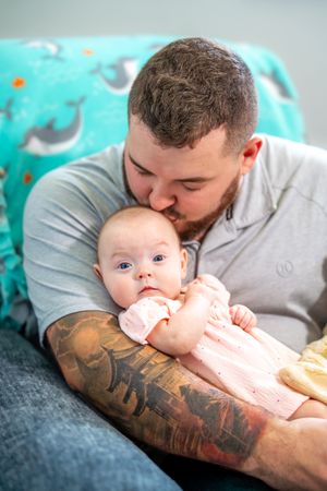 A father with a tattooed arm tenderly kisses his baby sitting on a couch with teal patterned cushions in the background. Quad Cities Newborn