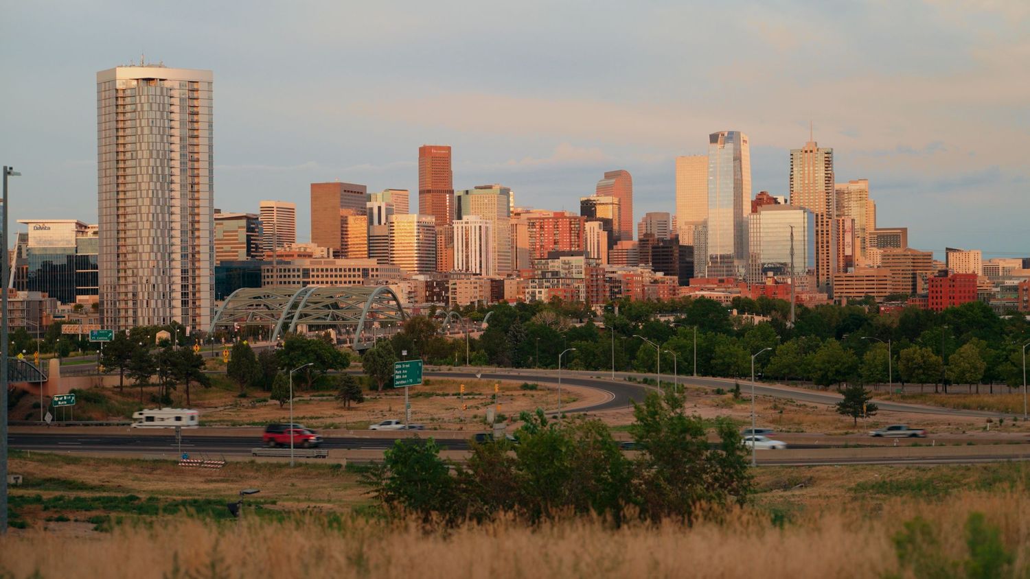 Downtown Denver skyline at sunset with high-rise buildings against an orange sky and urban park in the foreground.