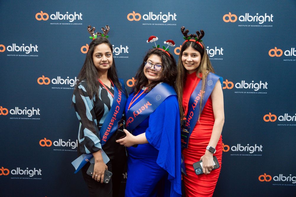 A row of graduates pose together in blue and red academic regalia against a branded backdrop at a ceremony.