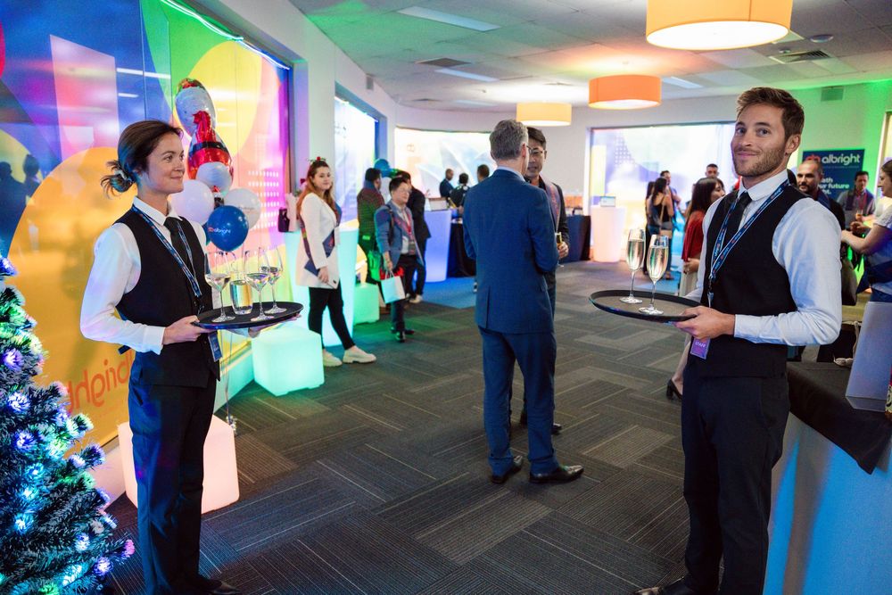 Waiters serving drinks at a colorful holiday party with festive lighting and a Christmas tree in the corner.