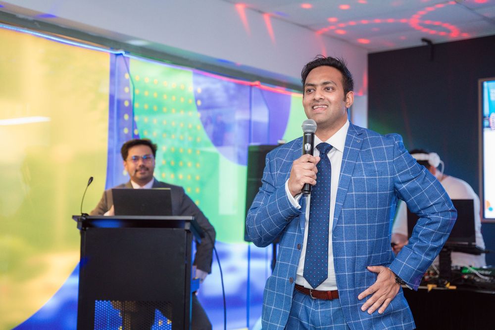 A presenter in a blue checkered suit jacket speaks on stage during a professional conference event.