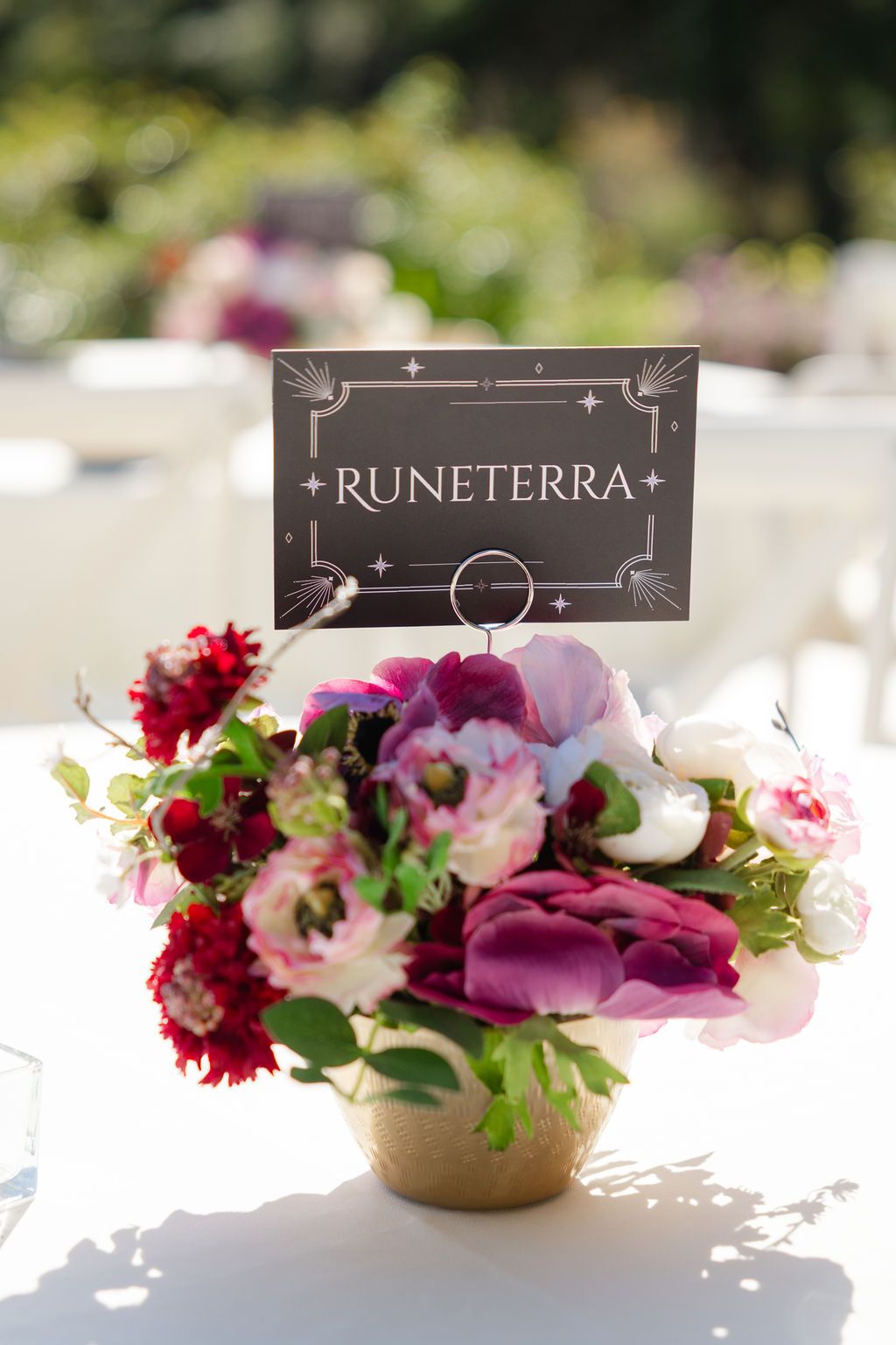 A floral centerpiece with pink and red flowers next to a black Eorzea sign in a gold vase on a white surface.